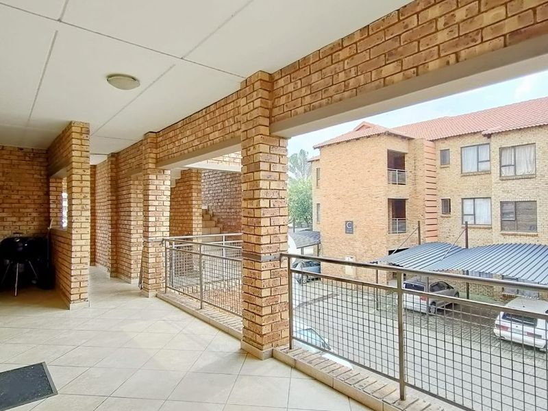 A spacious covered balcony featuring brick pillars, tiled flooring, and metal mesh railings, overlooking the apartment complex parking area.