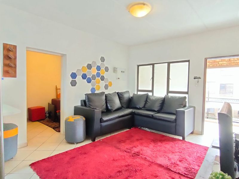 A living room featuring tiled flooring, aluminium window frames, and a ceiling light fixture, with direct access to an adjacent room and outdoor area.