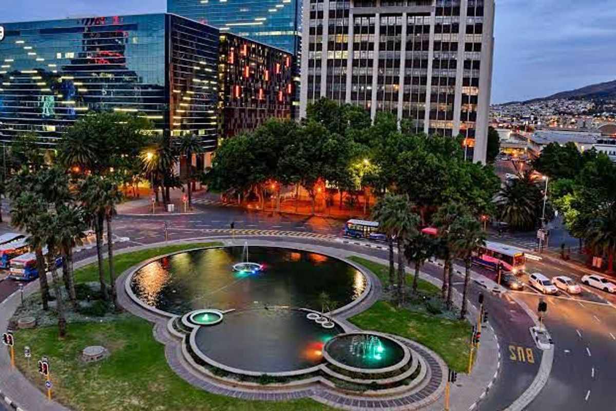 An aerial view of a landscaped urban roundabout featuring a multi-tiered fountain, surrounded by city buildings and traffic infrastructure.