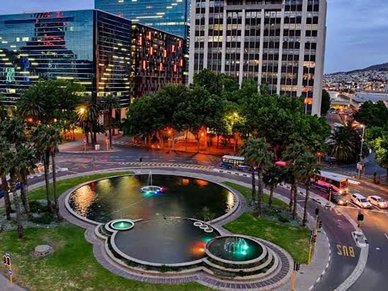 An aerial view of a landscaped urban roundabout featuring a multi-tiered fountain, surrounded by city buildings and traffic infrastructure.