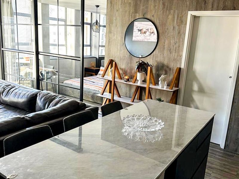 Modern open-plan living area featuring a sleek kitchen island with stone countertop, wood-look feature wall, and stylish black metal-framed glass room divider.