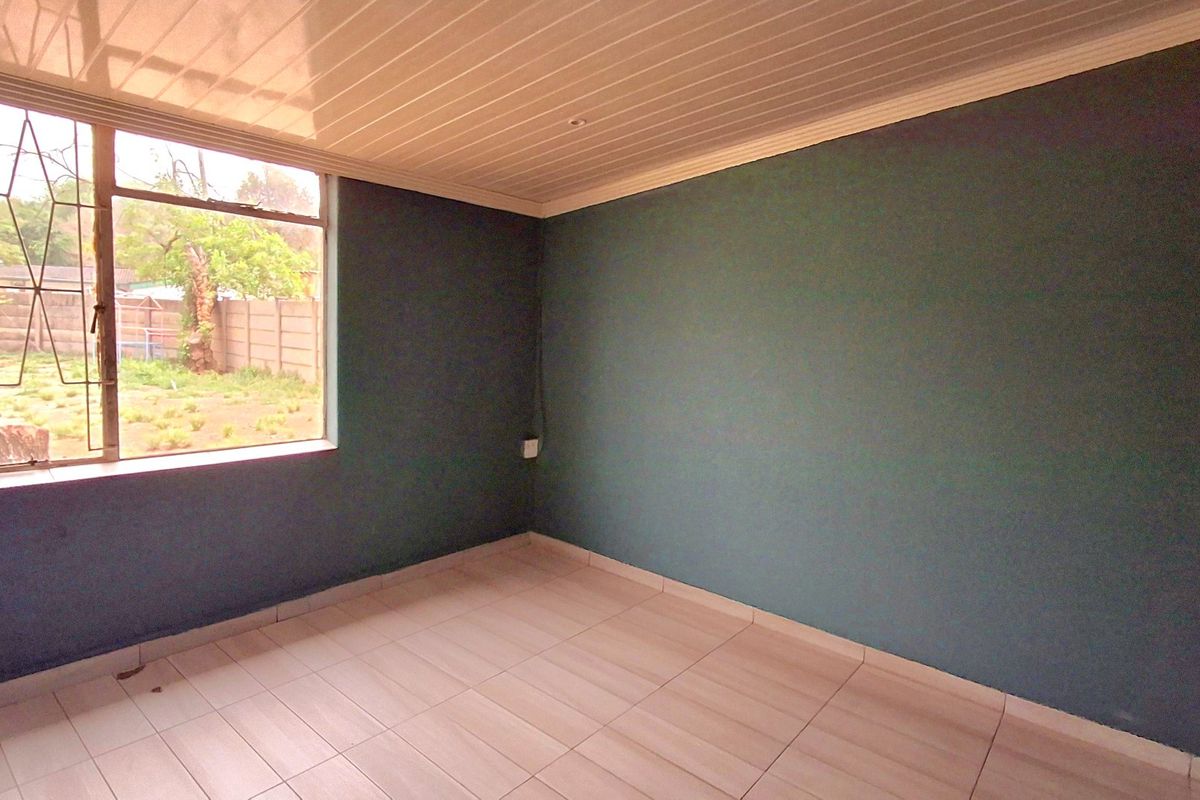 A bright, empty bedroom featuring tiled flooring, a large window with garden views, and a clean, neutral ceiling finish.