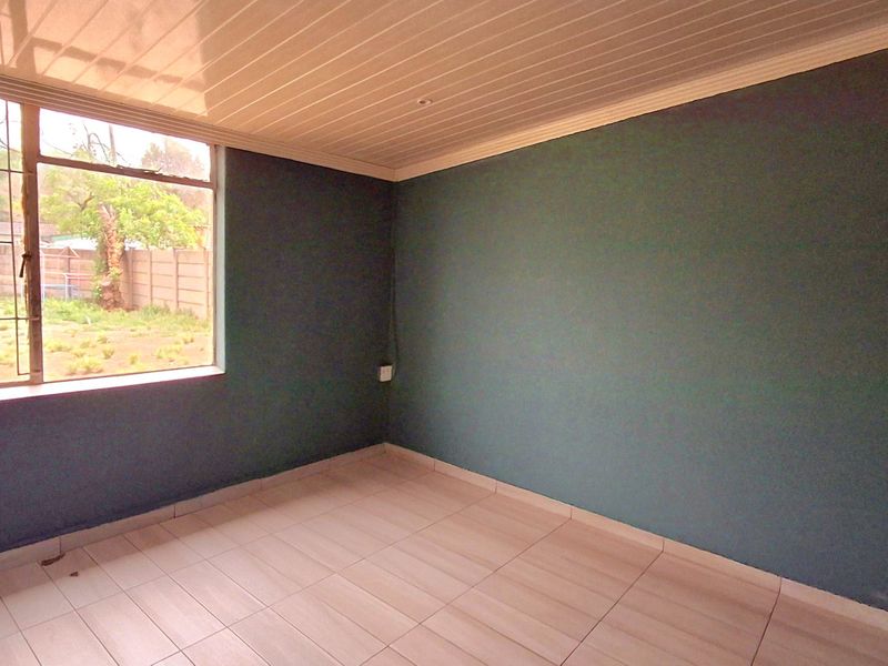 A bright, empty bedroom featuring tiled flooring, a large window with garden views, and a clean, neutral ceiling finish.