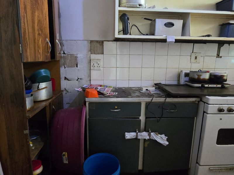 A dated kitchen area featuring built-in cabinetry, a tiled backsplash, and a freestanding stove, currently showing significant wear and missing wall tiles.
