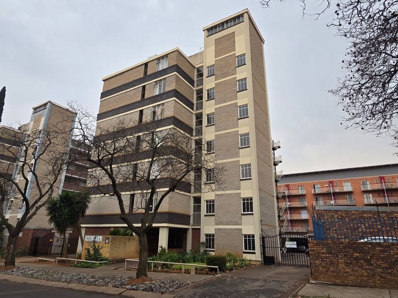 Exterior view of a multi-story apartment building featuring a brick facade, secure gated entrance, and paved driveway area.