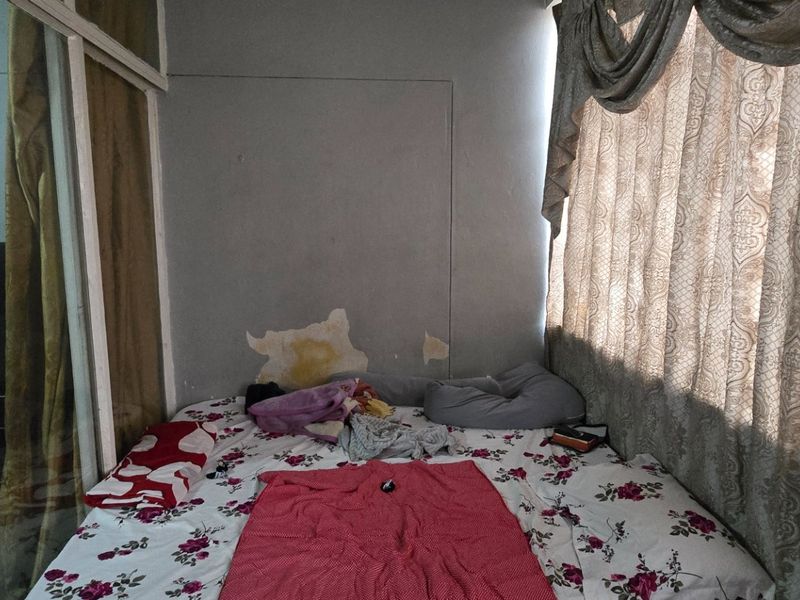A bedroom featuring a sliding glass partition and large windows with curtains, currently showing signs of wall surface deterioration.
