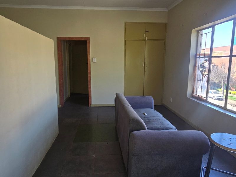 A living area featuring tiled flooring, a built-in cupboard, and large windows with security bars, offering a functional space in need of cosmetic updates.