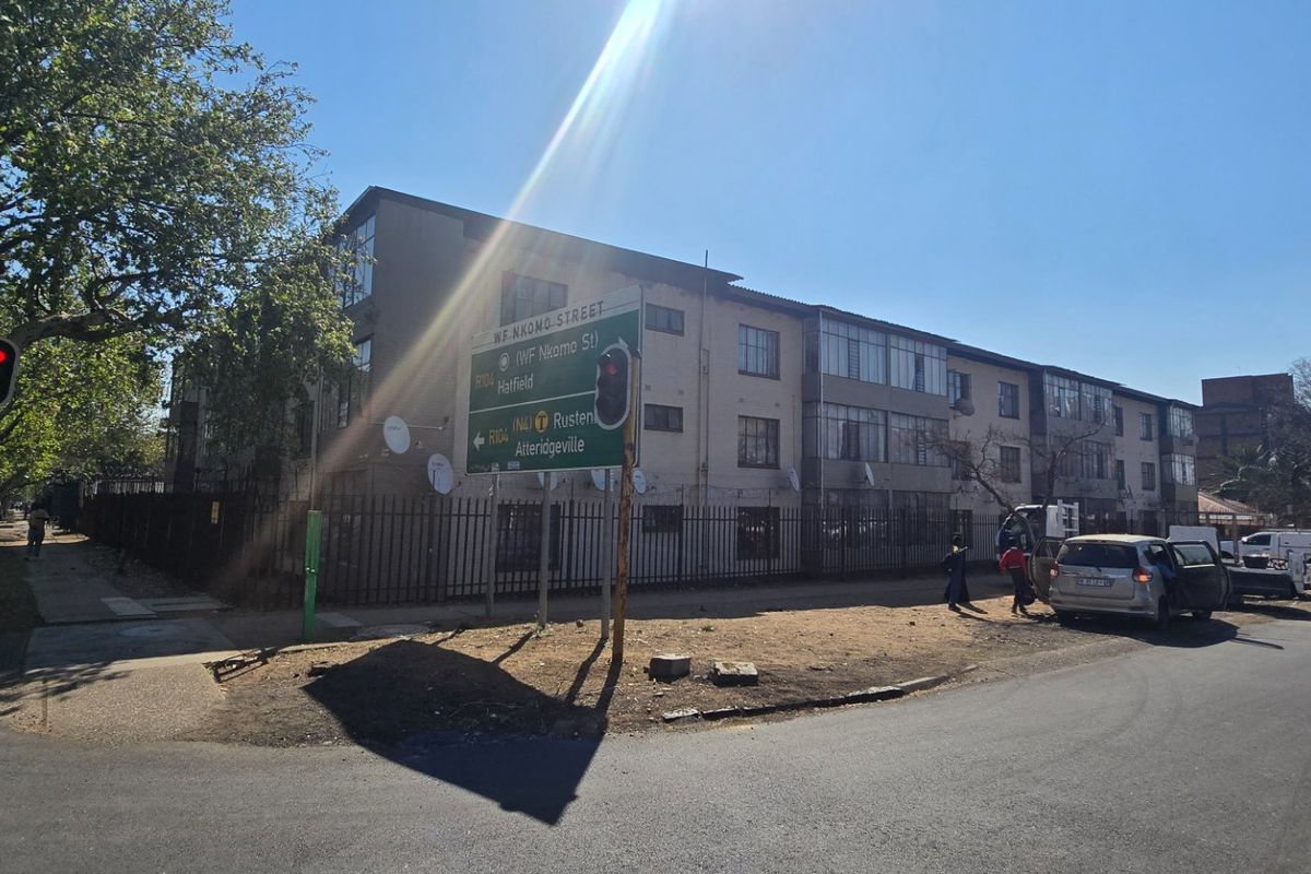 Street view of the apartment complex exterior, showing the building facade, security fencing, and surrounding road infrastructure.