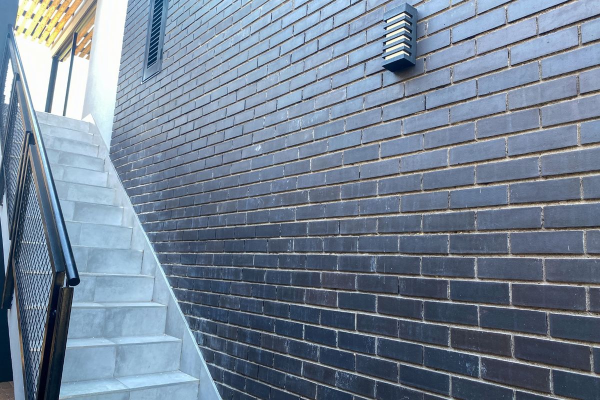 An exterior view of a modern apartment staircase featuring dark brickwork, a metal balustrade, and a timber pergola overhead.