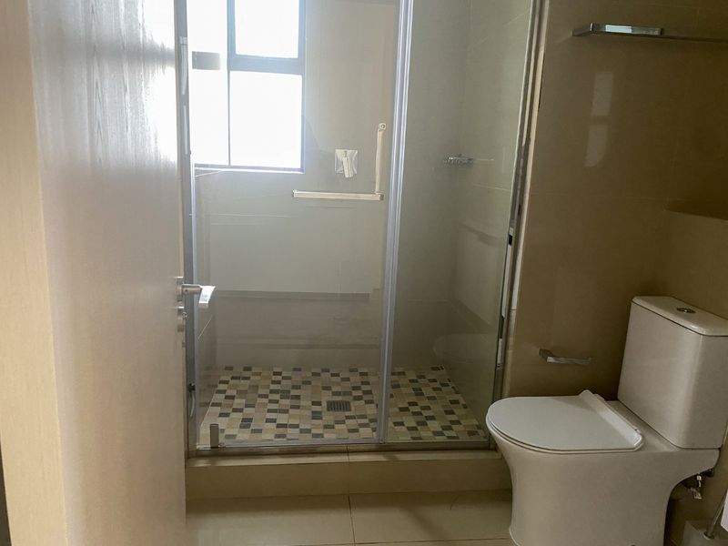 A functional bathroom featuring a glass-enclosed shower with mosaic floor tiling, a standard toilet, and neutral wall tiles.