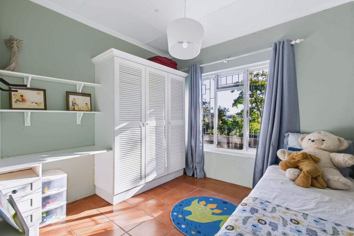 A bedroom featuring built-in cabinetry, a desk area, tiled flooring, and a window with security bars overlooking the garden.