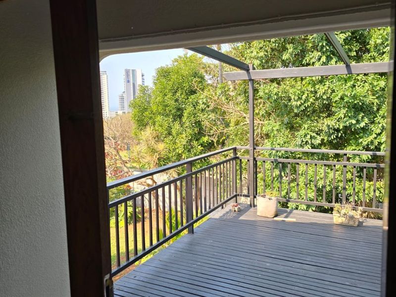 A covered balcony featuring timber decking, a metal balustrade, and a pergola structure, offering an elevated view of the surrounding trees and cityscape.