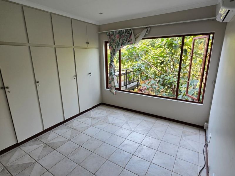 A bright, empty bedroom featuring tiled flooring, extensive built-in cupboards, recessed lighting, and a split-system air conditioner with a view of greenery.