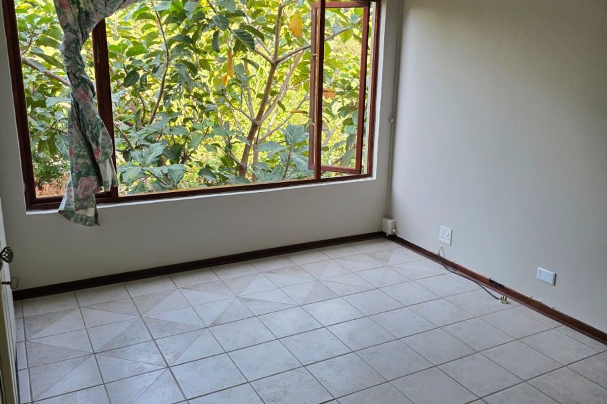 An empty bedroom featuring tiled flooring, recessed lighting, and a wall-mounted air conditioning unit with large windows overlooking greenery.