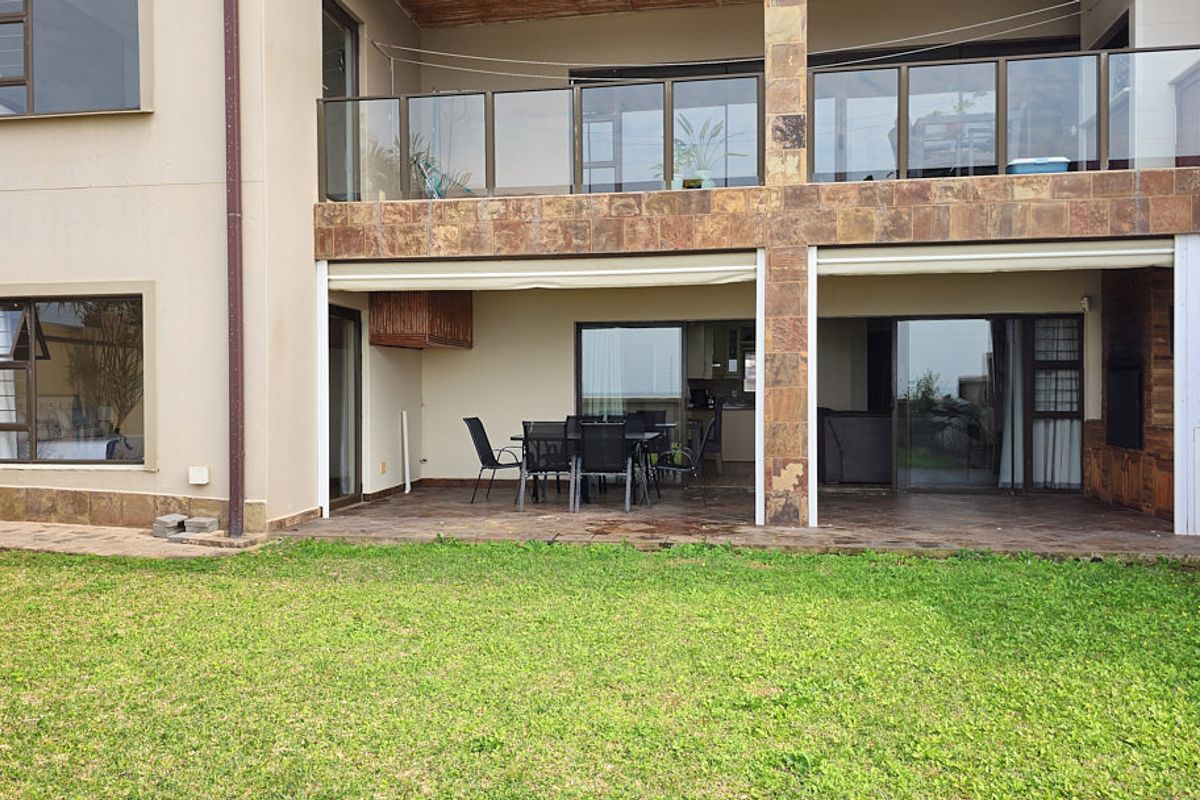 Rear view of the apartment featuring a covered ground-floor patio, an upper-level balcony with glass balustrades, and stone-clad structural pillars overlooking a garden.