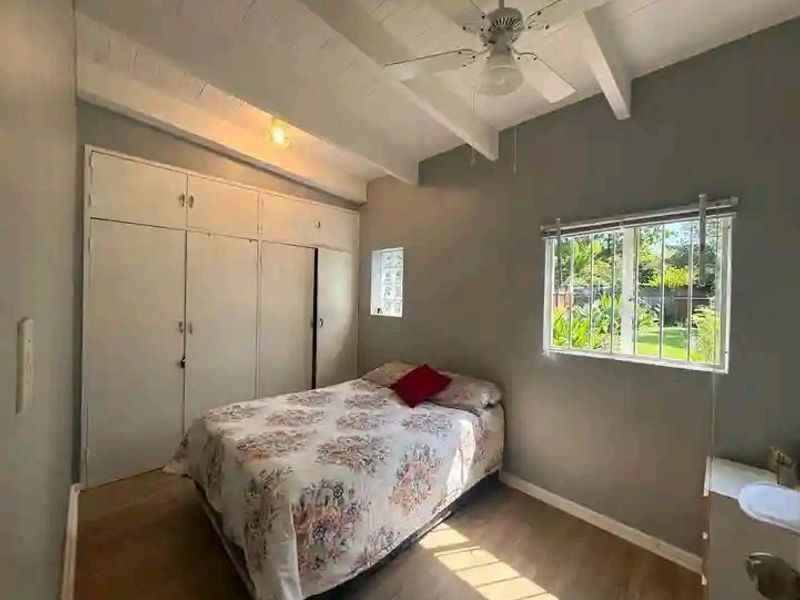 A bedroom featuring built-in cupboards, exposed beam ceiling with a fan, and a window with security bars overlooking the garden.