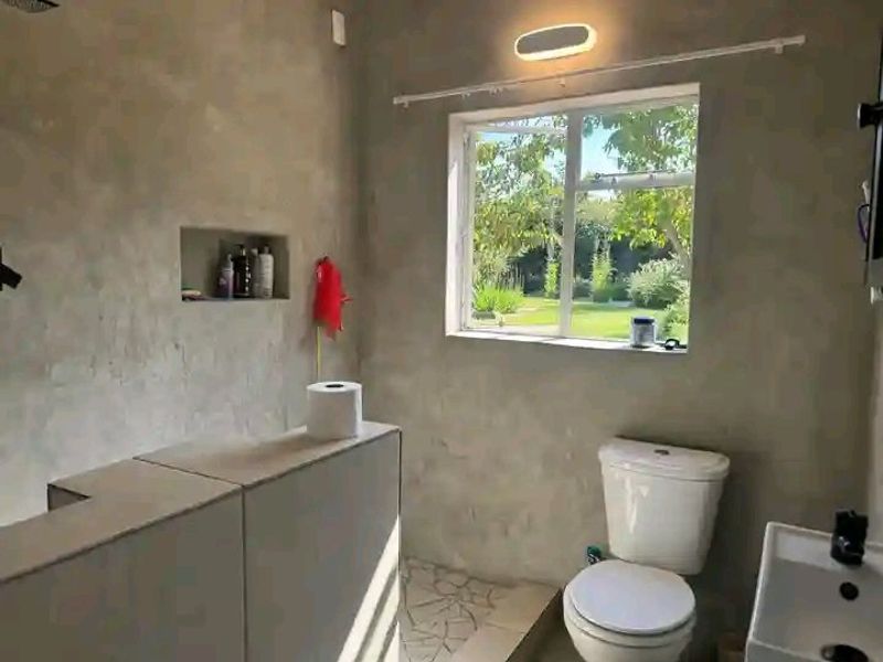 A bathroom featuring raw cement-finish walls, a built-in shower niche, a toilet, and a washbasin, with a window overlooking the garden.