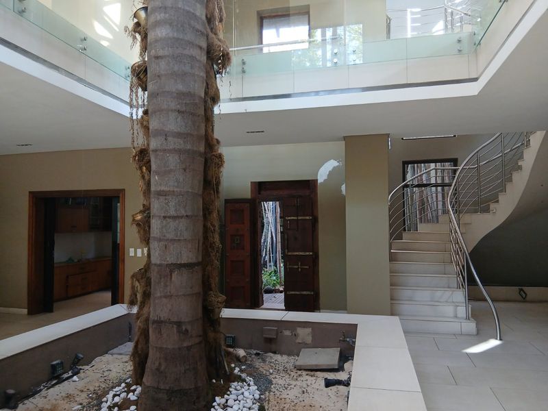 Spacious double-volume entrance hall featuring a central internal courtyard with a palm tree, a curved staircase, and modern glass and stainless steel balustrades.