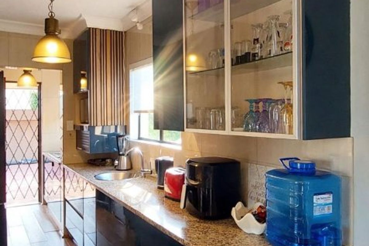 A modern galley-style kitchen featuring dark glossy cabinetry, granite countertops, glass-fronted display cabinets, and warm timber-look flooring.