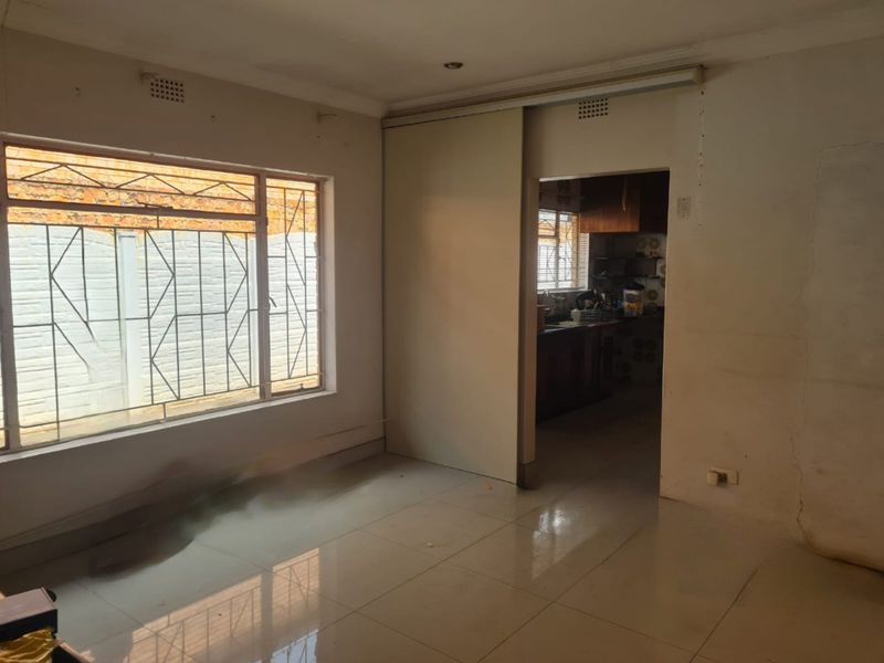 An empty living room featuring tiled flooring, a large window with security bars, and a sliding door leading to the kitchen area.