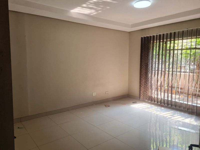 A bright, empty living room featuring neutral tiled flooring, recessed ceiling details, and large windows fitted with vertical blinds and security bars.