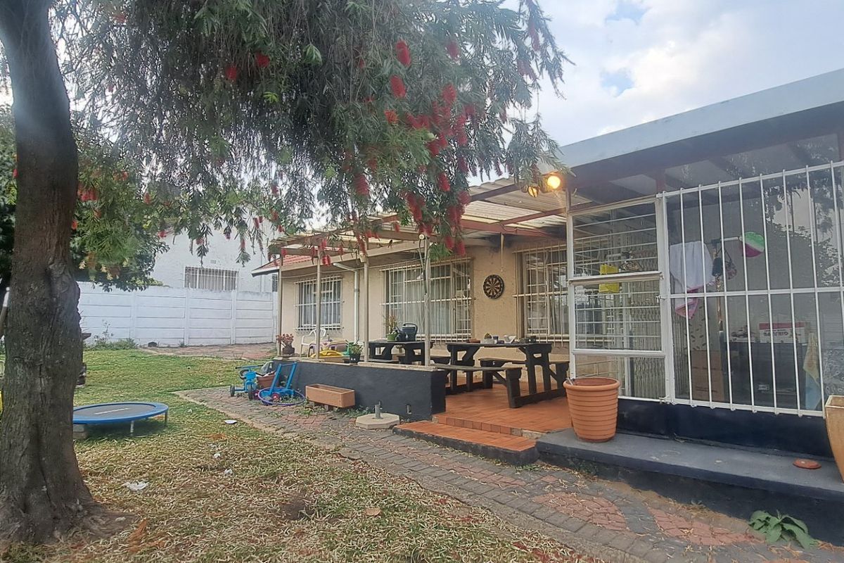Rear view of the property featuring a covered patio area, paved walkway, and a spacious garden with established trees and security features.