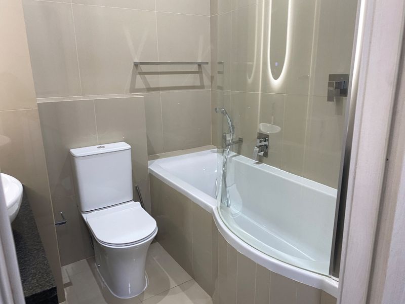 A modern, well-maintained bathroom featuring a built-in bathtub with a glass shower screen, wall-mounted toilet, and neutral tiled finishes.