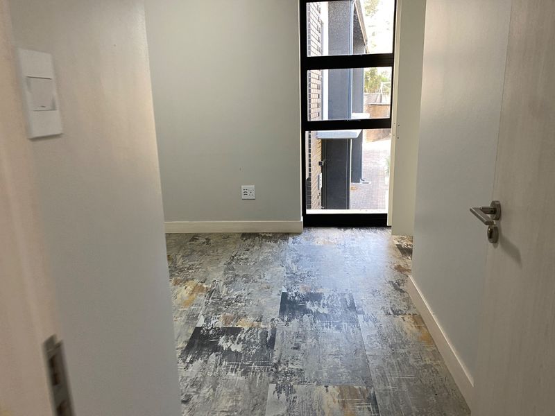 A bright, empty bedroom featuring modern patterned vinyl flooring, neutral walls, and a large black-framed window providing natural light.