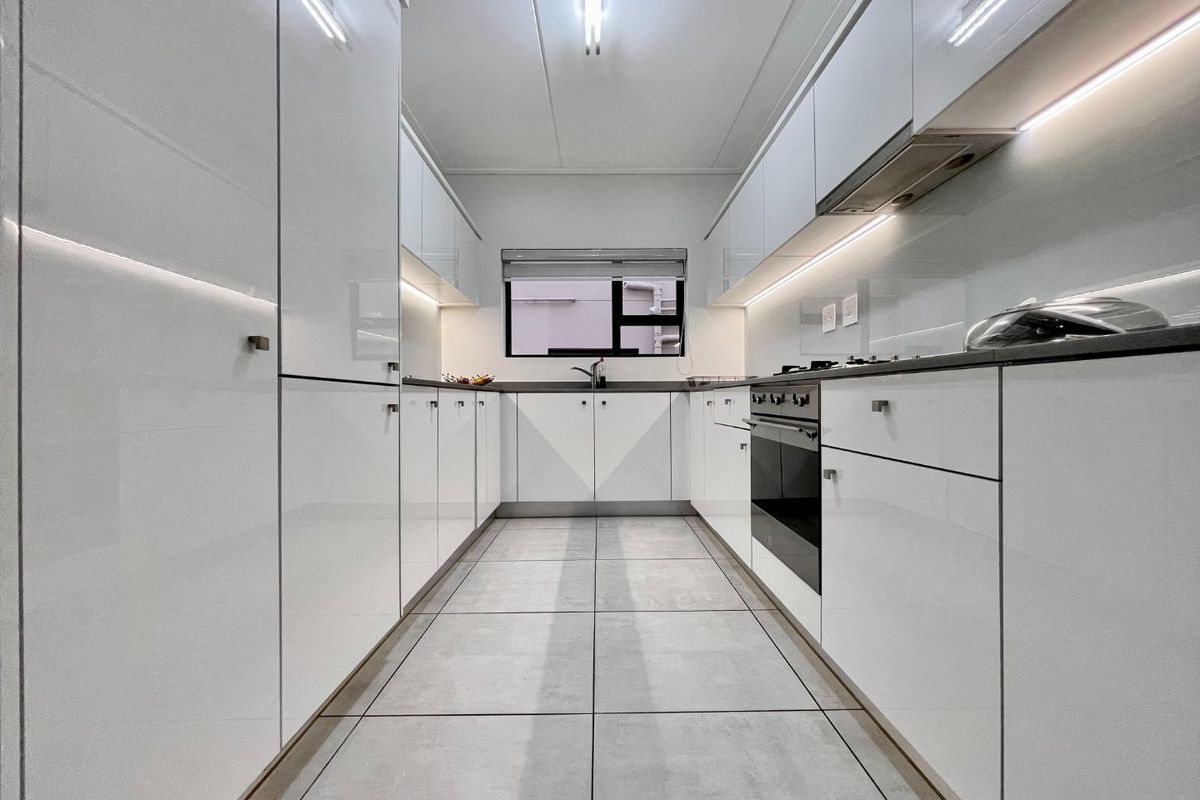 Modern galley-style kitchen featuring high-gloss white cabinetry, integrated appliances, under-cabinet lighting, and sleek tiled flooring.