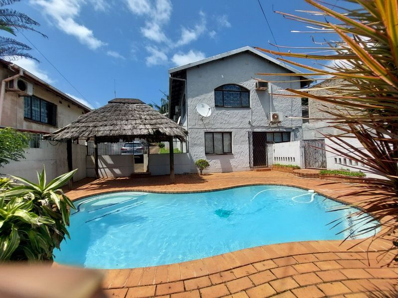 Rear exterior view featuring a swimming pool, brick paved patio area, and a traditional thatched lapa structure.