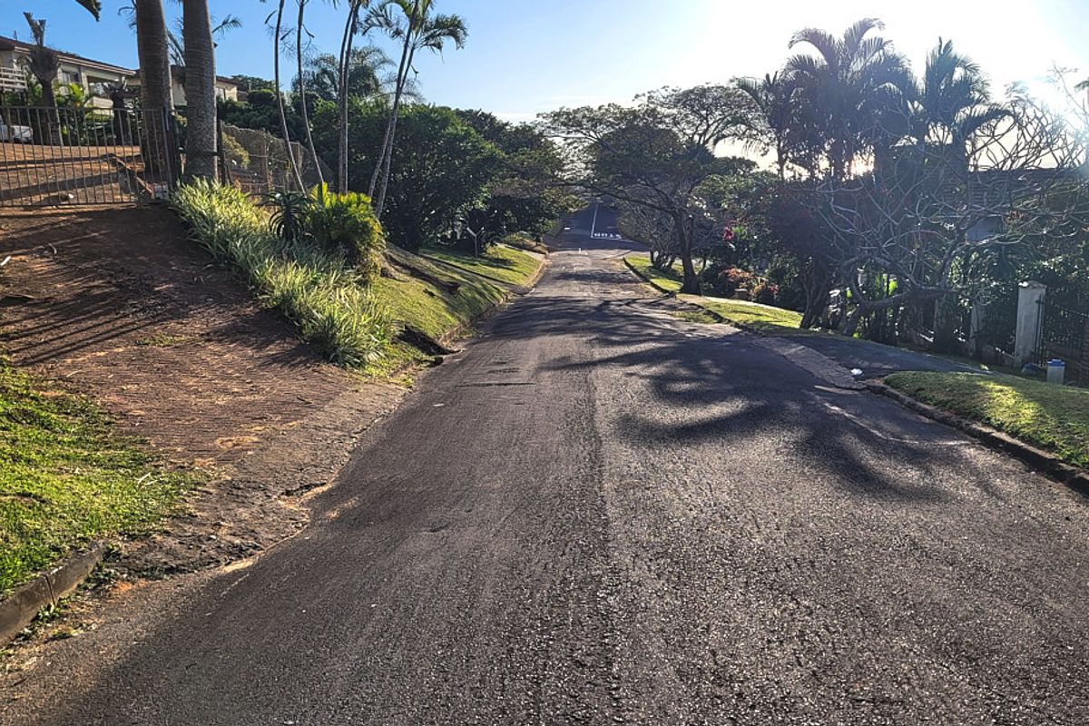 A view looking down a paved residential street with established trees, landscaped verges, and perimeter fencing along the property boundary.