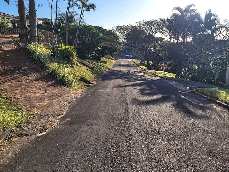 A view looking down a paved residential street with established trees, landscaped verges, and perimeter fencing along the property boundary.