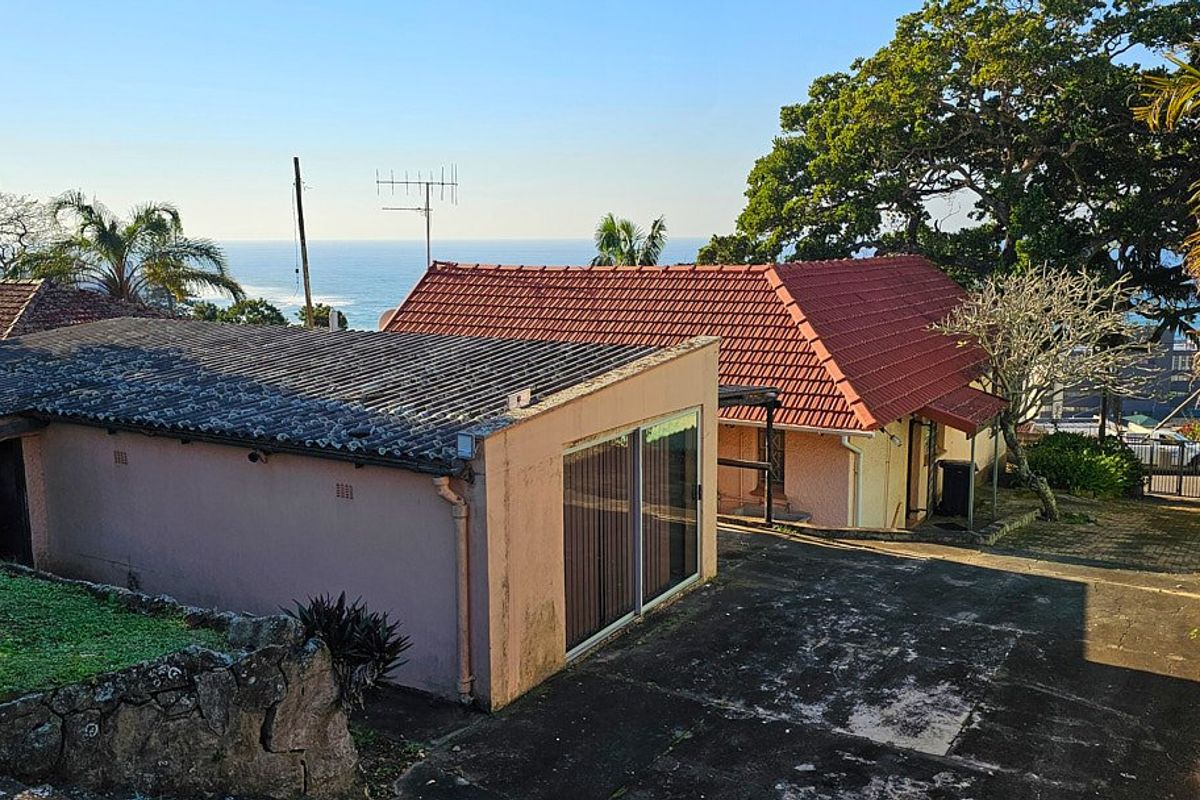 Rear view of the property featuring a paved driveway, stone retaining wall, and a combination of tiled and corrugated roofing with ocean views.