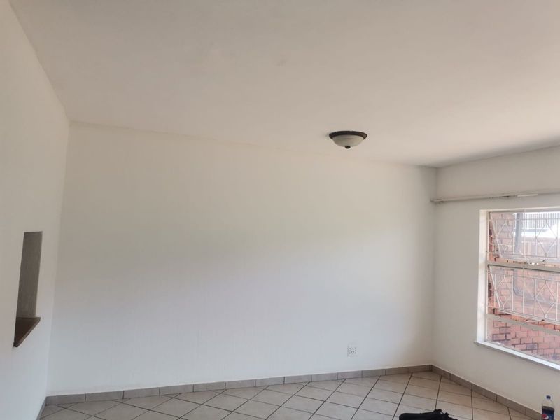 An empty living room featuring tiled flooring, a ceiling light fixture, and a window with security bars, offering a clean and neutral space.
