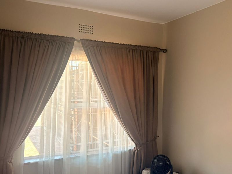 A simple bedroom corner featuring a window with curtains and a wall-mounted ventilation grille.