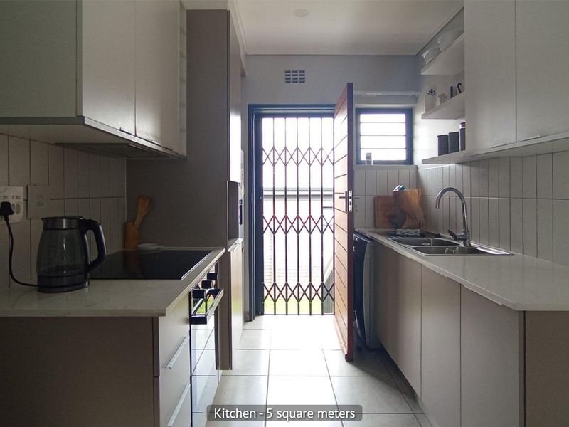 Compact galley-style kitchen featuring modern cabinetry, built-in oven and hob, stainless steel sink, and direct access to the exterior via a security gate.