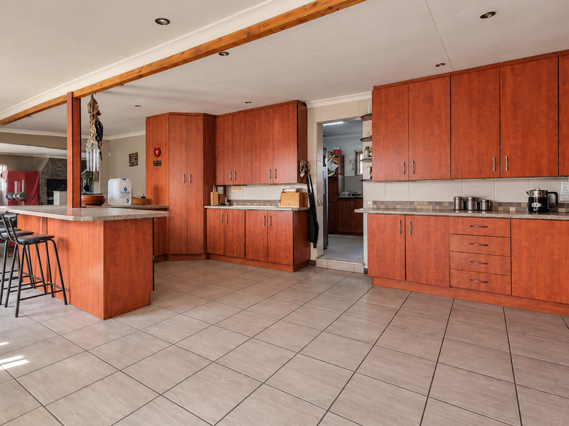 Spacious open-plan kitchen featuring warm-toned wooden cabinetry, stone countertops, a central island, and neutral tiled flooring, offering a functional and well-lit cooking space.