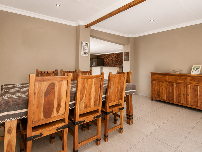 A well-maintained dining room featuring neutral wall tones, tiled flooring, and an exposed timber ceiling beam, with an open-plan flow to the kitchen area.