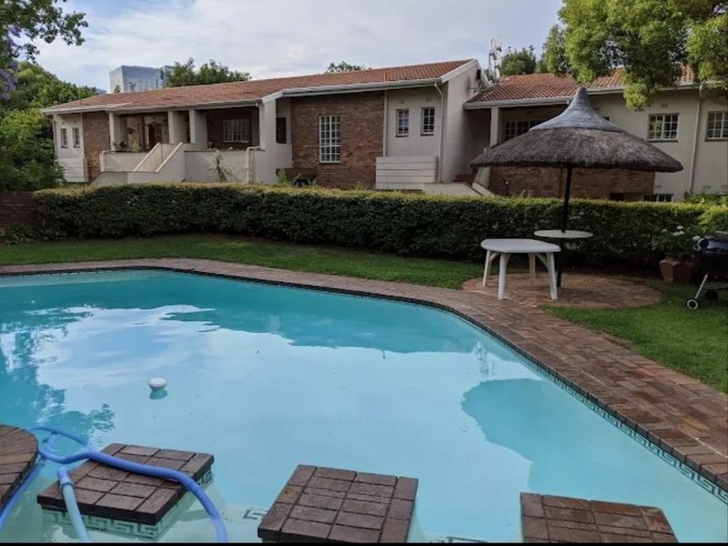 A private swimming pool area featuring brick paving and a thatched umbrella, set against the rear facade of a brick townhouse complex.