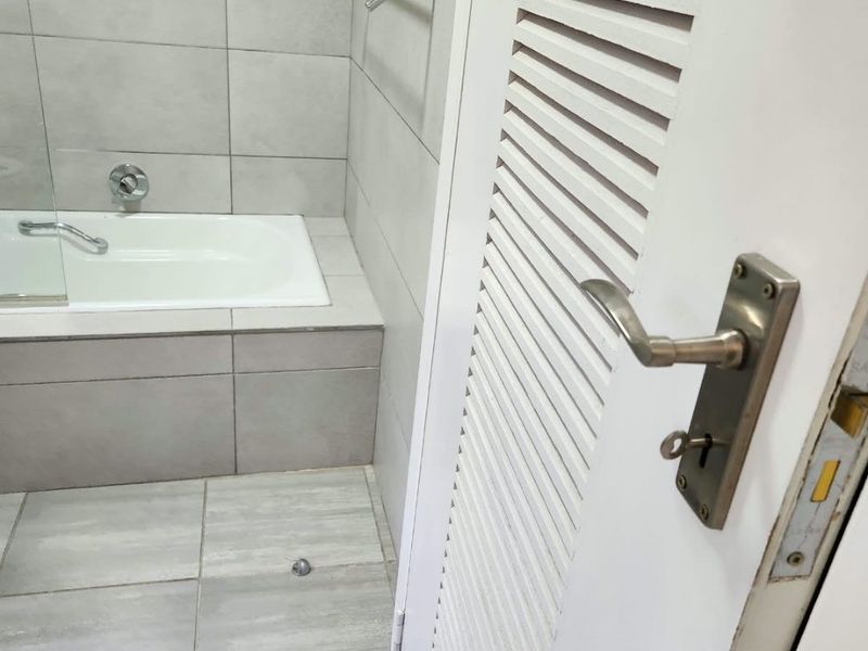 A functional bathroom featuring a built-in bathtub with a glass screen, neutral floor-to-ceiling tiling, and a louvered door.