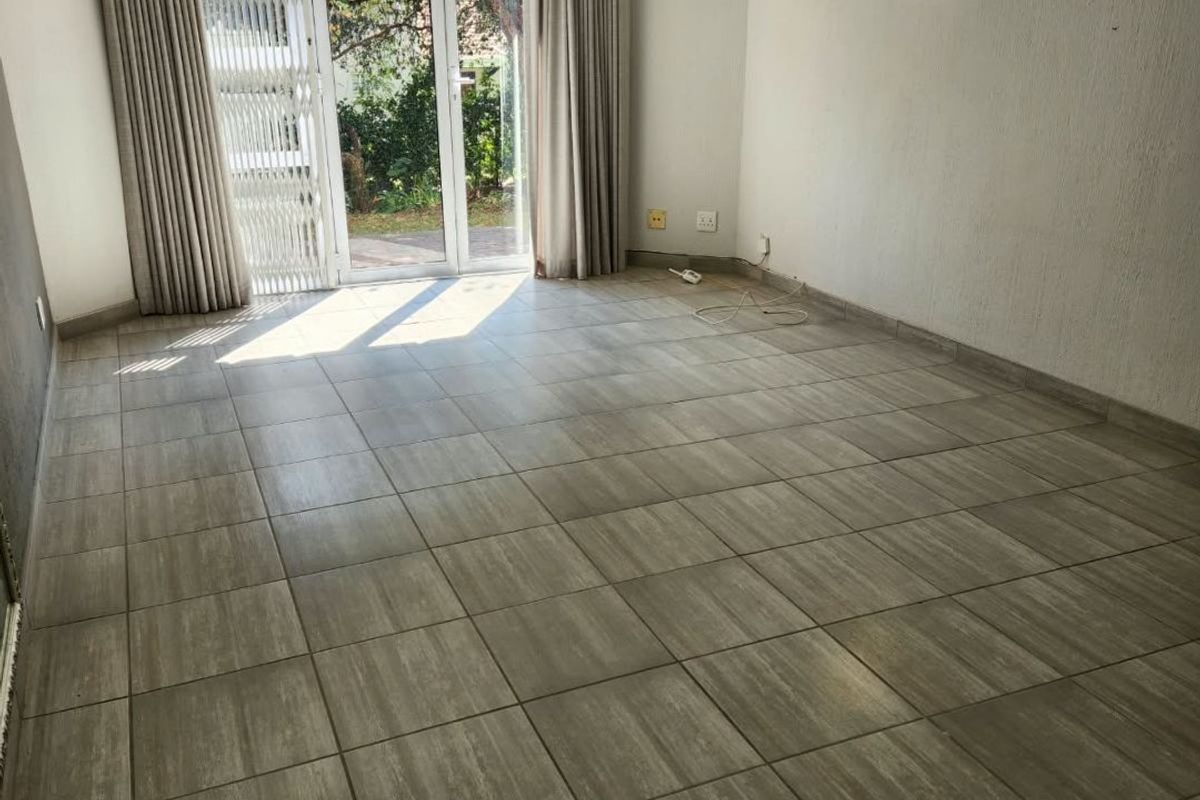 An empty living room featuring neutral tiled flooring, a sliding glass door with security gate, and direct access to the garden area.