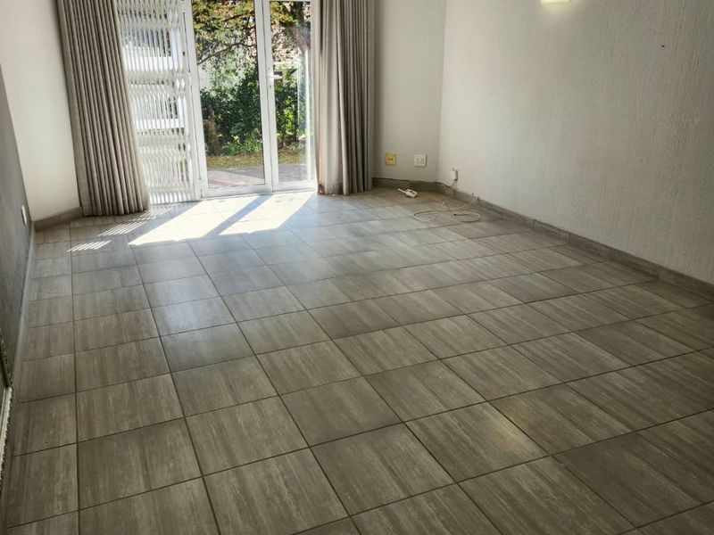 An empty living room featuring neutral tiled flooring, a sliding glass door with security gate, and direct access to the garden area.