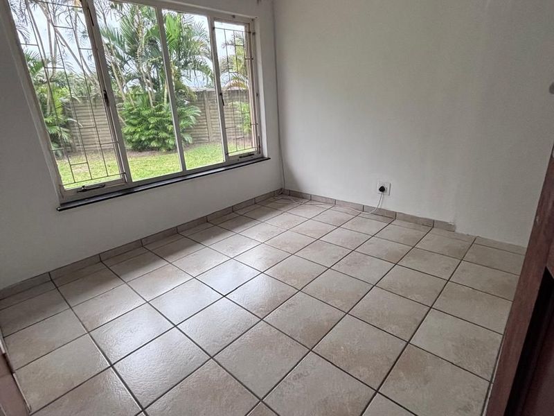 A bright, empty bedroom featuring neutral tiled flooring and a large window with security bars, offering a view of the garden.