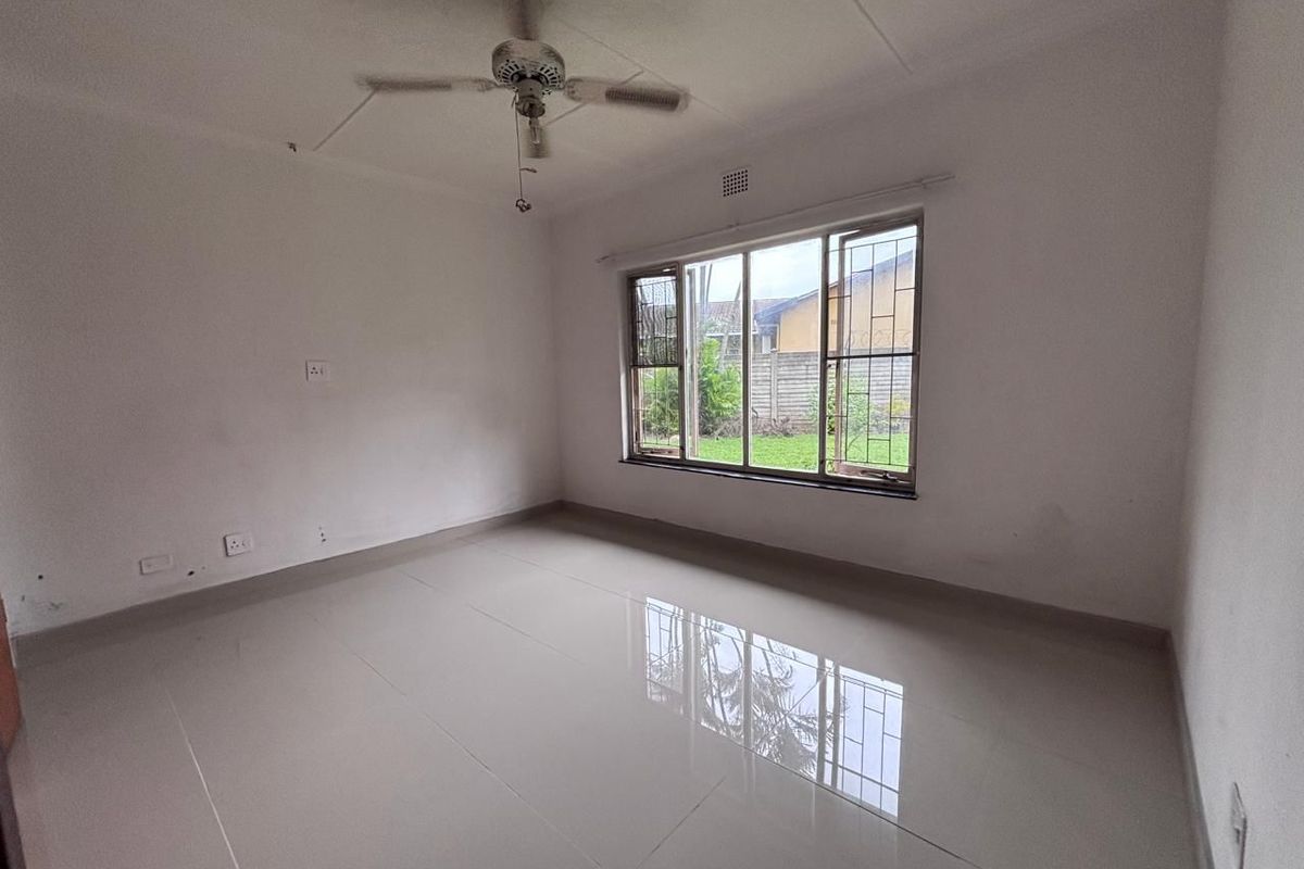 A vacant bedroom featuring neutral tiled flooring, a ceiling fan, and large windows with security bars overlooking the garden.