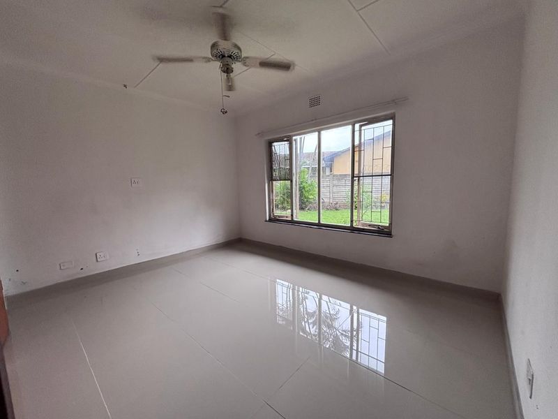 A vacant bedroom featuring neutral tiled flooring, a ceiling fan, and large windows with security bars overlooking the garden.