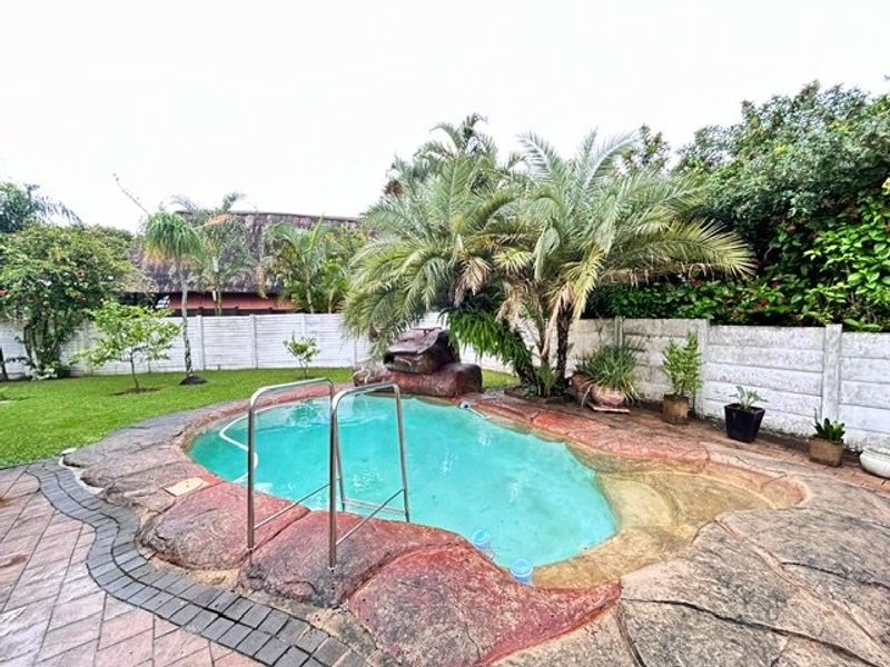 Outdoor pool area featuring a rock-style pool surround, metal ladder, paved patio, and a precast concrete boundary wall in a landscaped garden setting.
