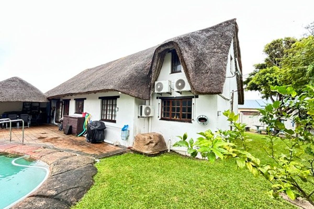 Rear exterior view of a thatched-roof property featuring a swimming pool, paved patio area, and established garden surroundings.
