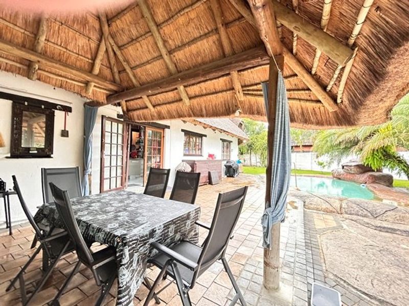 Covered outdoor patio area featuring a traditional thatched roof, timber support posts, and paved flooring, overlooking a private swimming pool.