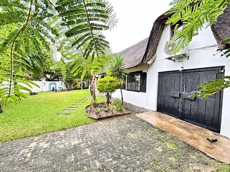 Rear view of a property featuring a thatched roof, paved driveway, and a well-maintained garden area with boundary wall.