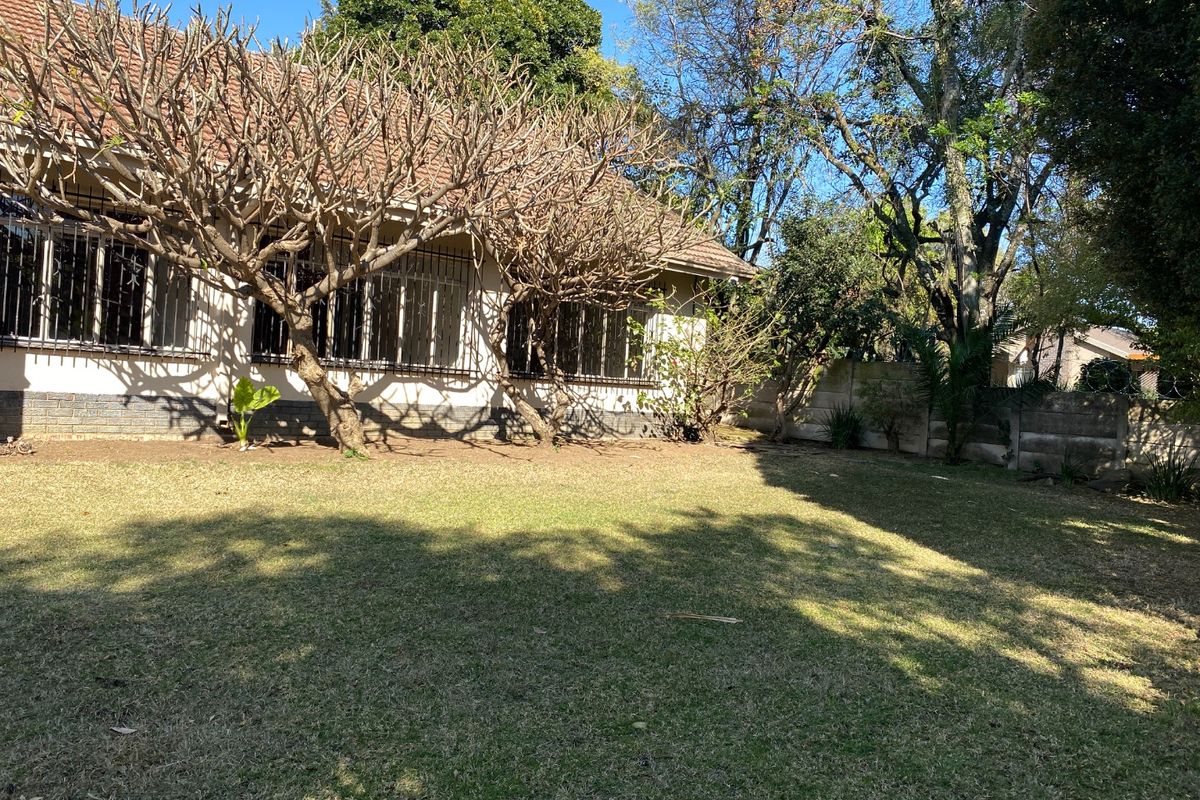 A view of the rear garden featuring a lawn area, mature trees, and the back elevation of the house with security bars on windows.